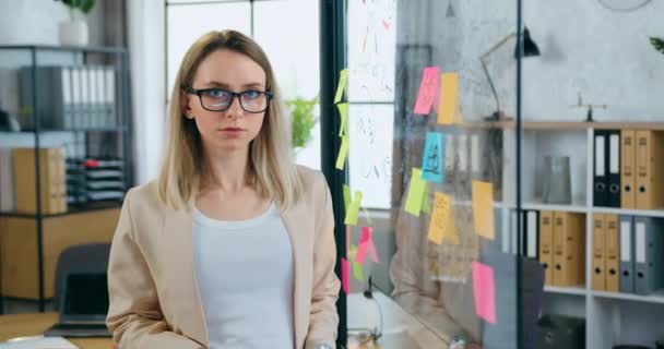 Belle souriante femme blonde qualifiée avec succès debout près du conseil transparent dans le bureau moderne et regardant dans la caméra, portrait de femme d'affaires — Video