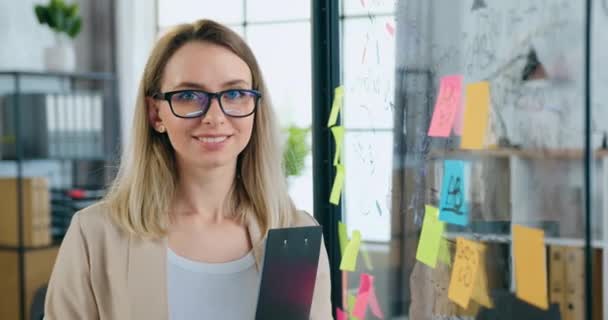 Retrato femenino de encantadora sonriente hábil mujer successul en gafas que mirando a la cámara con la cara feliz mientras está de pie con la carpeta cerca de tablero de vidrio con notas en pegatinas y tablero — Vídeo de stock