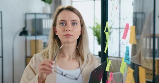 Nahaufnahme einer ziemlich seriösen erfahrenen 30-jährigen blonden Frau, die in der Nähe ihres Arbeitsplatzes in die Kamera blickt, mit einer Glastafel mit wichtigen Geschäftsdaten auf Aufklebern, die daran erinnern — Stockvideo