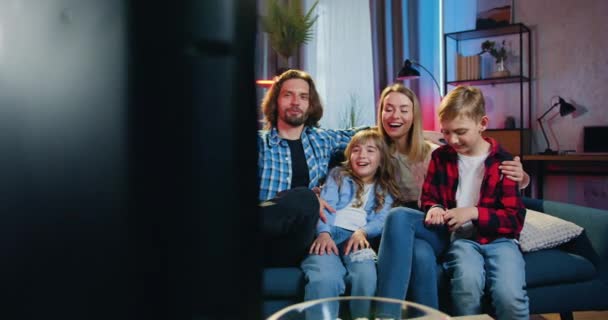 Portret van een schattig, vrolijk en positief modern gezin dat 's avonds op de bank rust en samen plezier beleeft terwijl we naar een interessante show op tv kijken — Stockvideo