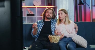 Aile atmosferi konsepti. Çekici, kaygısız bir çift TV karşısında oturmuş patlamış mısır yiyor, konuşuyorlar ve iki çocuk arkalarından atlayıp koltukta zıplıyorlar.