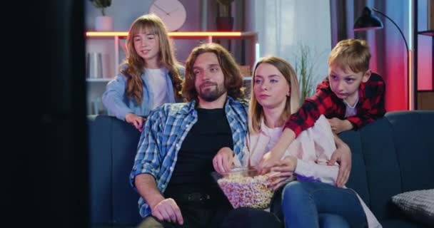 Familje idyll där attraktiva glada leende föräldrar och deras två barn tittar på TV-show hemma på kvällen och äter popcorn från glasskål — Stockvideo