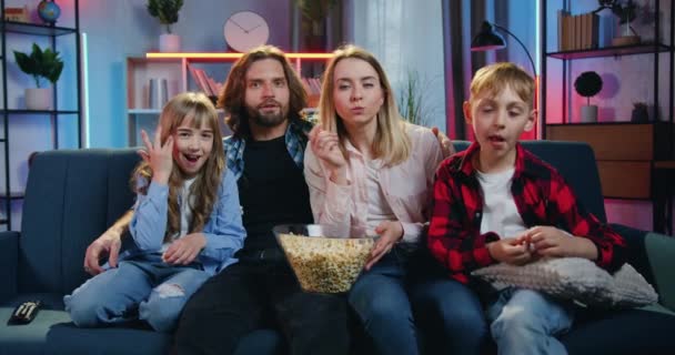 Portret van een heerlijk vrolijk glimlachend gezin van vier personen - moeder, vader, zoon en dochter die 's avonds thuis op de bank zitten, tv kijken en popcorn proeven — Stockvideo