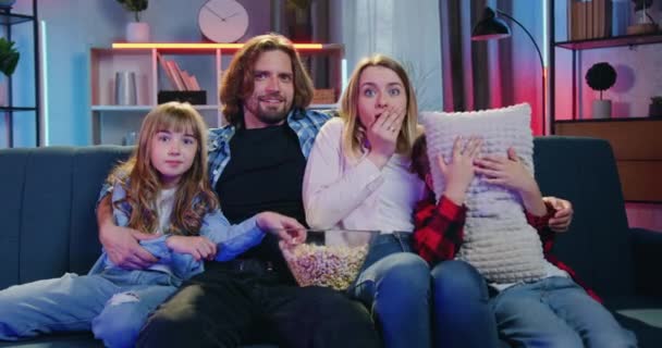 Rodzinny wypoczynek, gdzie atrakcyjne przestraszonych rodziców z dziećmi siedzi razem na kanapie i ogląda horror w telewizji w domu wieczorem — Wideo stockowe