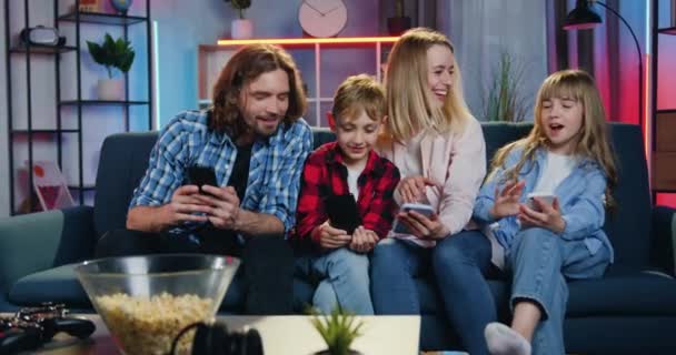 Atraktivní šťastně se usmívající moderní rodina čtyř osob - matka, otec, syn a dcera sedí doma na pohovce a používají své telefony k chatu na sociálních sítích nebo prohlížení fotografií