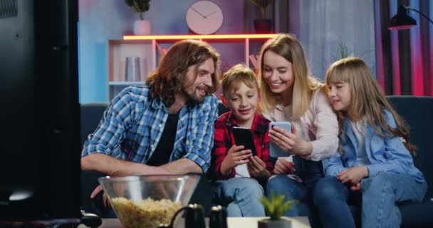 Atraktivní úsměv šťastný milující rodina odpočívá na pohodlné pohovce doma ve večerních hodinách a rodiče prohlížení fotografií na svých dětí mobily