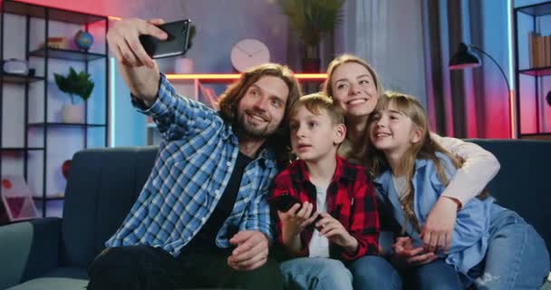 Adorable szczęśliwy uśmiechnięty przyjazny rodzina siedzi razem na kanapie w domu w godzinach wieczornych i robi selfie na smartfonie, nowoczesnej technologii i koncepcji wypoczynku rodzinnego — Wideo stockowe