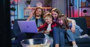 Güzel görünüşlü mutlu modern ailenin portresi. Akşamları evdeki kanepede oturup dizüstü bilgisayardaki eğlenceli videoların keyfini çıkarıyorlar.