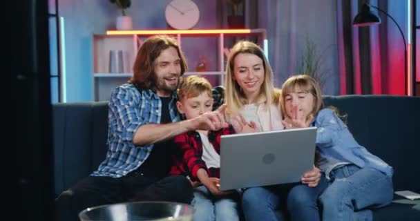 Agradable sonriente joven familia satisfecha sentada en el sofá suave en casa por la noche durante el chat de vídeo con familiares en el ordenador portátil y saludarlos con las manos agitadas — Vídeos de Stock