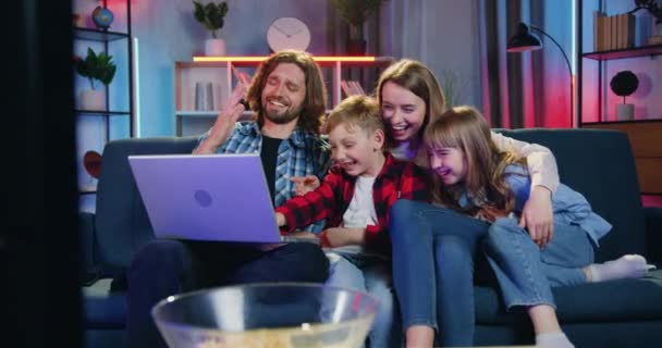 Retrato de boa aparência alegre família moderna feliz que sentado no sofá em casa à noite e desfrutando de rever vídeos conjuntos engraçados no laptop — Vídeo de Stock