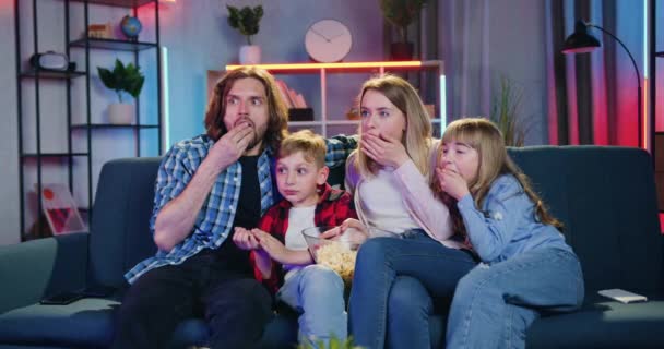 Portrait of attractive positive surprised family which watching breathtaking film on TV at home in the evening simultaneously eating popcorn,leisure concept — Stock Video