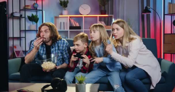 Casa conceito de entretenimento familiar onde bonito emocional duas crianças jogando videogames na TV set em casa à noite, juntamente com seus pais animados engraçados que apoiá-los e se alegrar de — Vídeo de Stock