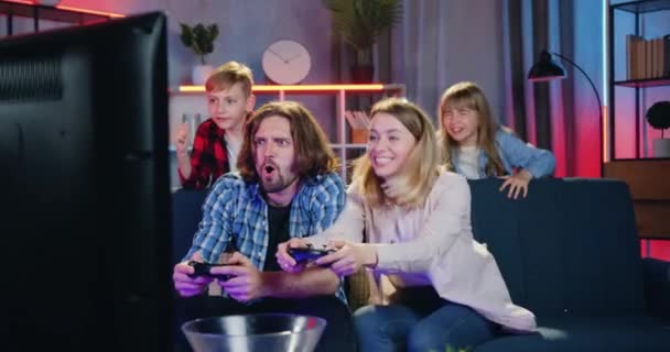 Χαριτωμένο χαμογελαστά συναισθηματικά παιδιά που υποστηρίζουν ενθουσιασμένοι παιχνιδιάρικο αστείο γονείς τους που παίζουν βιντεοπαιχνίδια στην τηλεόραση στο σπίτι το βράδυ, μπροστινή άποψη — Αρχείο Βίντεο