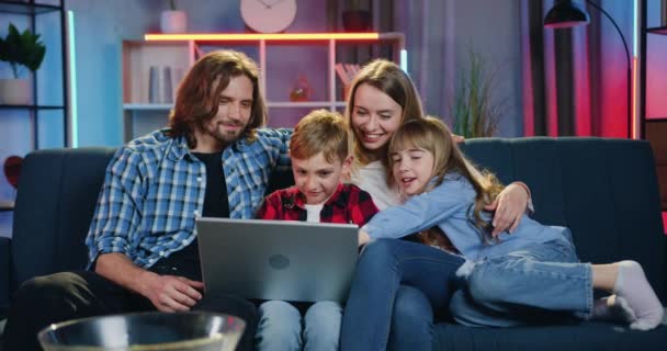 Primer plano de la atractiva feliz y sonriente pareja amorosa con dos hijos que revisionan las fotos familiares en el ordenador mientras pasan el ocio conjunto en casa por la noche — Vídeo de stock