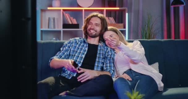 Preciosa feliz sonriente joven pareja enamorada sentados juntos en un sofá suave en la sala de estar por la noche y ver la comedia en la televisión, la mujer pone su cabeza en el hombro novios — Vídeo de stock