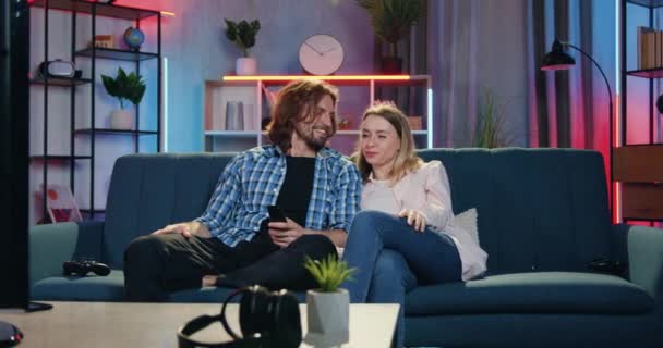 Boa aparência feliz sorrindo casal jovem amoroso conversando entre si enquanto assiste filme engraçado na TV no sofá macio no quarto aconchegante à noite, conceito de lazer — Vídeo de Stock