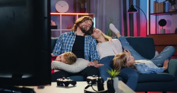 Heerlijk vermoeide familie met twee kinderen die 's avonds op de bank voor de werkende tv slapen, kinderen leggen hun hoofd op de knieën van hun ouders, familie relaxatie concept — Stockvideo
