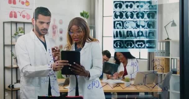 ガラスの壁の近くで働く化学者の魅力的な熟練した多様な医療グループタブレットPCからの情報とDNAコードの異なる化学式、科学の概念 — ストック動画
