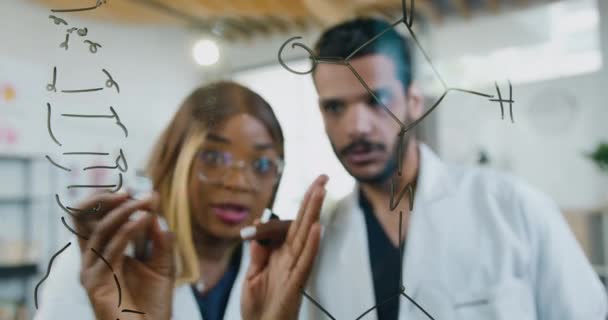 Genetik hastalıklardan gelen iki tıbbi araştırmacı üzerinde kimyasal elementlerin moleküler formülünü yazan cam duvarın yanında duruyor. — Stok video