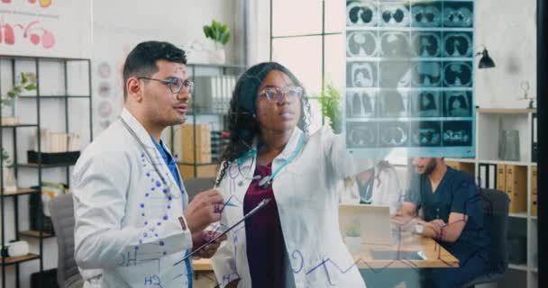 Snygga högkvalificerade självsäkra olika manliga och kvinnliga läkare undersöker resultaten av patienter röntgen fäst vid glasväggen i läkarmottagningen — Stockvideo