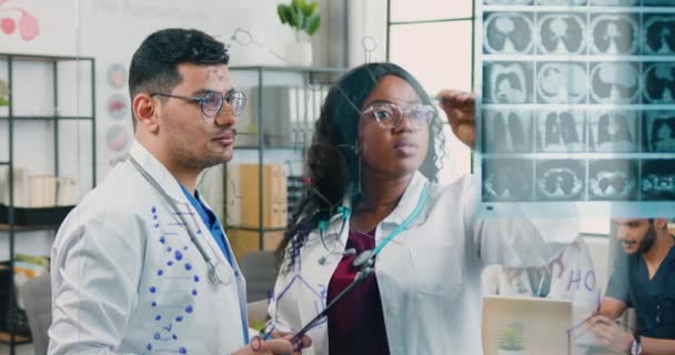 Sympathisch geschickte erwachsene gemischte Rasse männliche und weibliche Ärzte diskutieren Röntgenbild an der Glaswand in der Arztpraxis angebracht und die Wahl der Behandlung für Patienten — Stockvideo