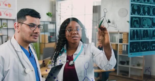 Close up of good-looking purposeful adult multiracial two medical researchers which analyzing chemical formula on glass wall in lab during joint work over methods of treatment of diseases — Stock Video