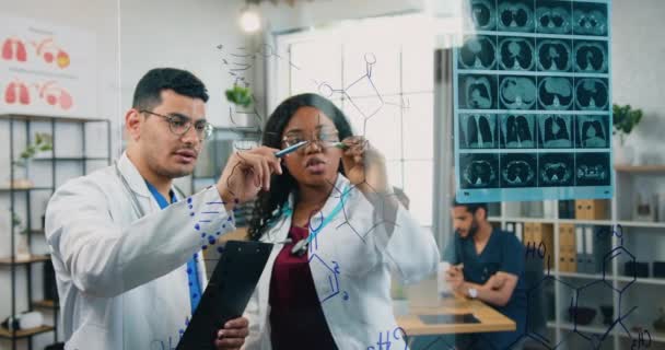 Вид через стеклянную доску, где уверенно высококвалифицированная смешанная раса двух мужчин и женщин химиков, которые работают со структурными формулами нарисованы на этой доске — стоковое видео