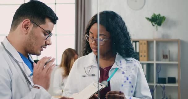 30 yaşında güzel bir profesyonel afro-amerikan kadın doktor, tıp ofisinde yetenekli sakallı erkek meslektaşıyla konuşuyor ve hasta tedavi yöntemlerini tartışıyor. — Stok video