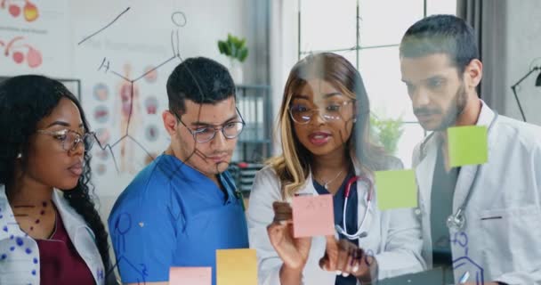 Multiracial team of skillful chemists working together over scientific formula and male colleague of group writing structural formula on glass board in lab — Stock Video