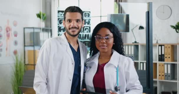 Портрет привлекательных улыбающихся профессиональных высококвалифицированных врачей смешанной расы, которые стоят перед камерой в медицинском кабинете в современной больнице — стоковое видео