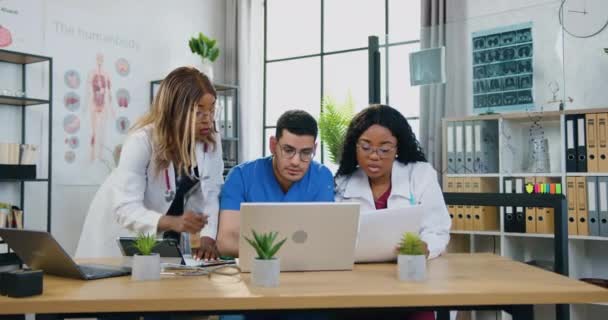 Atractivo seguro de sí mismo experimentado adulto de raza mixta tres médicos en uniforme trabajando juntos en la sala médica utilizando la información de la computadora y el informe de paer — Vídeo de stock