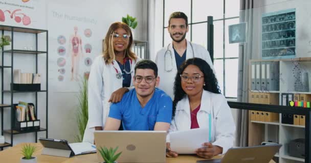 Портрет доброзичливих досвідчених позитивних лікарів змішаної раси, які позують у камеру в сучасній медичній кімнаті — стокове відео