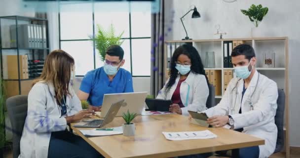 Squadra medica mista seria e sicura di sé in maschere protettive brainstorming insieme nella sala di consultazione in ospedale, lavoro durante la pandemia covid-19 — Video Stock