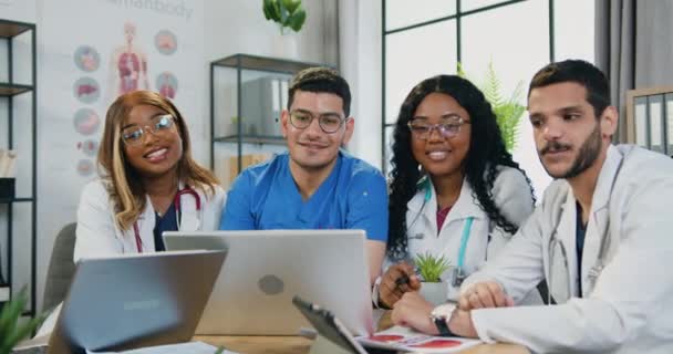 Portrait d'attrayant souriant professionnel hautement qualifié multiracial médical personnel qui est assis autour du lieu de travail dans la salle médicale et geste symbole bien dans la caméra — Video