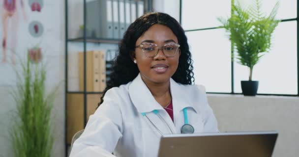 Gros plan de charmante professionnelle souriante cvalifiée femme médecin à la peau noire à l'aube blanche qui est assis devant la caméra avec ordinateur et symbole gestuel d'accord — Video