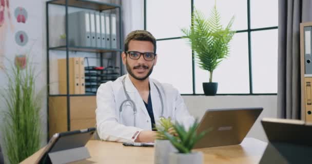 Piacevole gioioso fiducioso medico maschio barbuto cvalified in occhiali e camice bianco seduto sul suo posto di lavoro con il computer e guardando la fotocamera con sorriso sincero — Video Stock