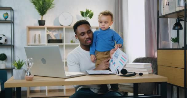 Remote werk en kinderopvang concept waar goed uitziende zelfverzekerde liefdevolle Afrikaanse Amerikaan houdt zijn kleine zoon op de knieën tijdens de uitleg van het verslag met statistieken door middel van video chat thuis — Stockvideo