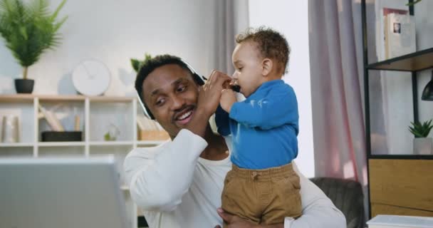 Concept de travail à distance et de garde d'enfants où le beau papa habile et souriant à la peau noire parle sur le chat vidéo avec ses collègues lorsque son petit enfant joue avec un microphone attaché aux pères — Video