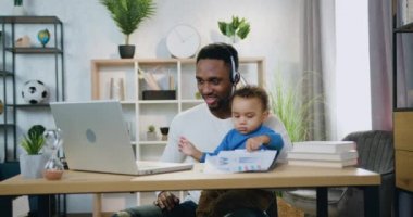 Yakışıklı, gülümseyen, umursamaz, siyah derili, kulaklıklı bir babanın portresi. Küçük oğlunu dizlerinin üstünde tutuyordu. Evde dizüstü bilgisayar başında, çocuk bakımı konseptinde.