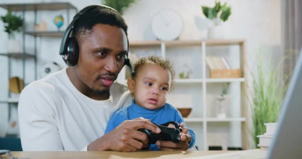 Närbild porträtt av stilig omtänksam positiv aktiv svarthyade far som spelar videospel på datorn tillsammans med sin söta lilla son hemma — Stockvideo