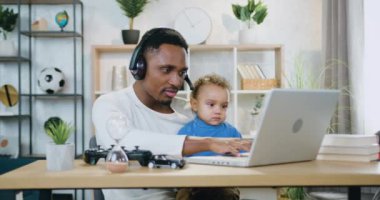 Evde bilgisayar, uzaktan çalışma ve çocuk bakımı konseptiyle uğraşırken küçük oğlunu dizlerinin üzerine çöktüren, kulaklık takan çekici, iyi niyetli, siyah tenli adam.