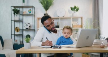 Sempatik, genç, siyah tenli bir babanın cep telefonuyla konuşması, kucağında bebek varken not alması, iş yerinde oturması, uzak mesai saatlerinde çocuk bakımı.
