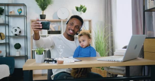 Dobře vypadající usmívající šťastný moderní černošky-kůže otec ve sluchátkách dělat selfie spolu se svým malým synem doma — Stock video