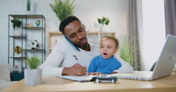 Bonito sorrindo carinhoso jovem pai de pele preta falando ao telefone, fazendo anotações em caderno e assistindo a seu pequeno filho que bocejando porque quer dormir, conceito de paternidade — Vídeo de Stock
