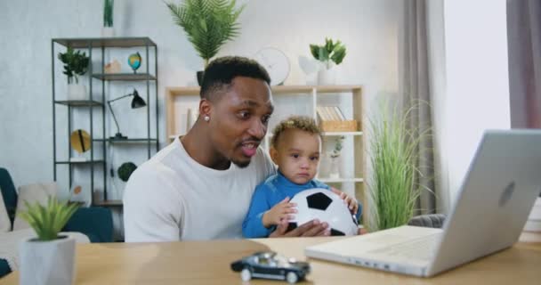 Jeu probable émotionnel attentionné père à la peau noire tenant son bébé garçon calme sur les genoux pendant le match de football de regarder sur ordinateur et criant après but marqué — Video