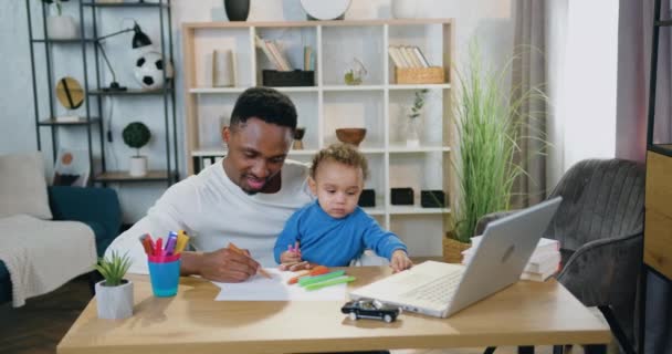 Péče o děti koncept, kde dobře vypadající šťastný uvolněný starostlivý mladý černoch kreslení spolu se svým roztomilým chlapečkem s barevnými plstěnými pery doma, přední pohled — Stock video
