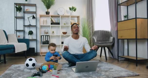 Aantrekkelijke kalme jonge zwarte huid man mediteren op de vloer met gesloten ogen in de buurt van zijn schattige kleine zoon die speelt met veel speelgoed, kinderopvang en recreatie concept — Stockvideo