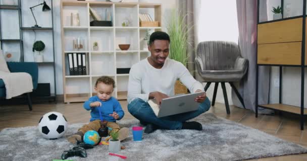 Aangename positieve bekwame zorgzame zwart-huid vader zitten op de vloer en werken aan laptop, terwijl zijn schattige kalme baby jongen spelen in zijn buurt met speelgoed, kinderopvang en remote werk concept — Stockvideo