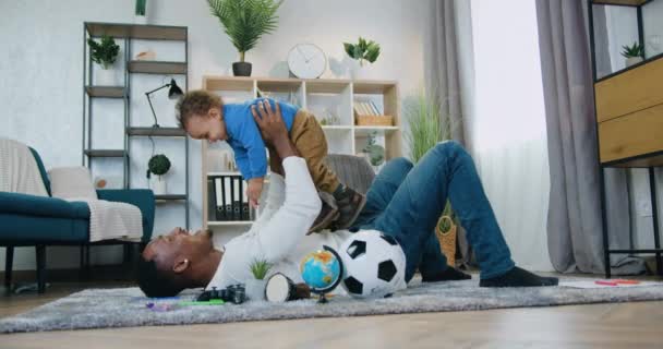 Jóképű gondtalan boldog pozitív fekete bőrű apa fekszik a földön otthon, és felemeli az aranyos vidám kisfiú a kezét, gyermekgondozás koncepció — Stock videók