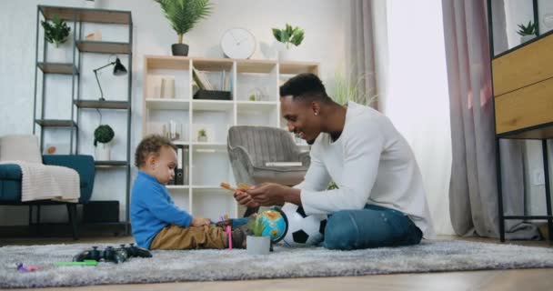 Dobrze wyglądający szczęśliwy uśmiechnięty, troskliwy, afrykański ojciec pomaga zebrać długopisy dla swojego słodkiego synka, gdy bawią się razem w domu. — Wideo stockowe